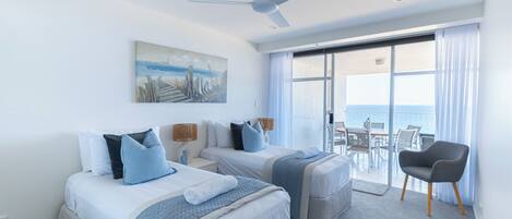 A hotel room with two beds, a ceiling fan, a painting, a balcony with a view of the sea, and a chair.