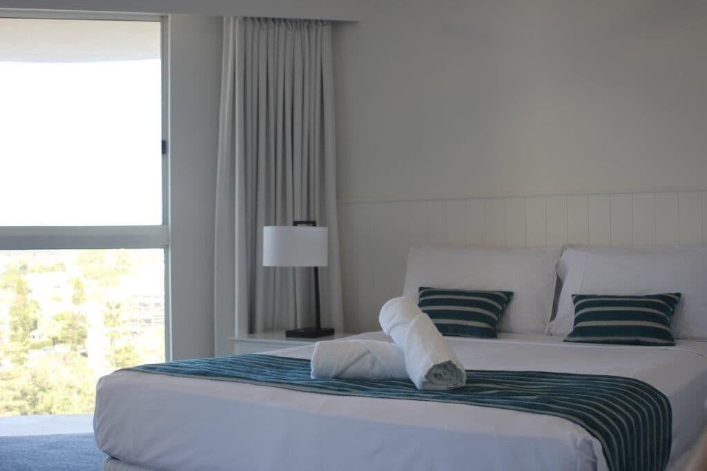 Ein Hotelzimmer mit großem Fenster, einem Bett mit weißer Bettwäsche und gestreiften Kissen, einem Nachttisch mit Lampe und Blick auf Bäume.