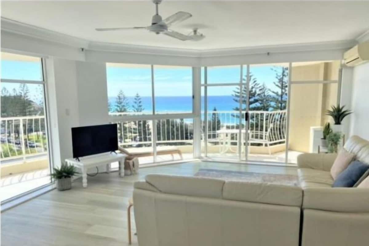 A spacious living room with a large window offering a view of the ocean and trees, a ceiling fan, and a flat-screen TV on a stand.