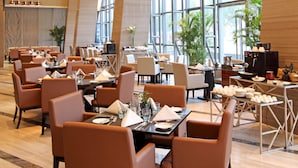 Een ruime hotel eetzaal met meerdere tafels en stoelen, grote ramen en moderne verlichtingsarmaturen.