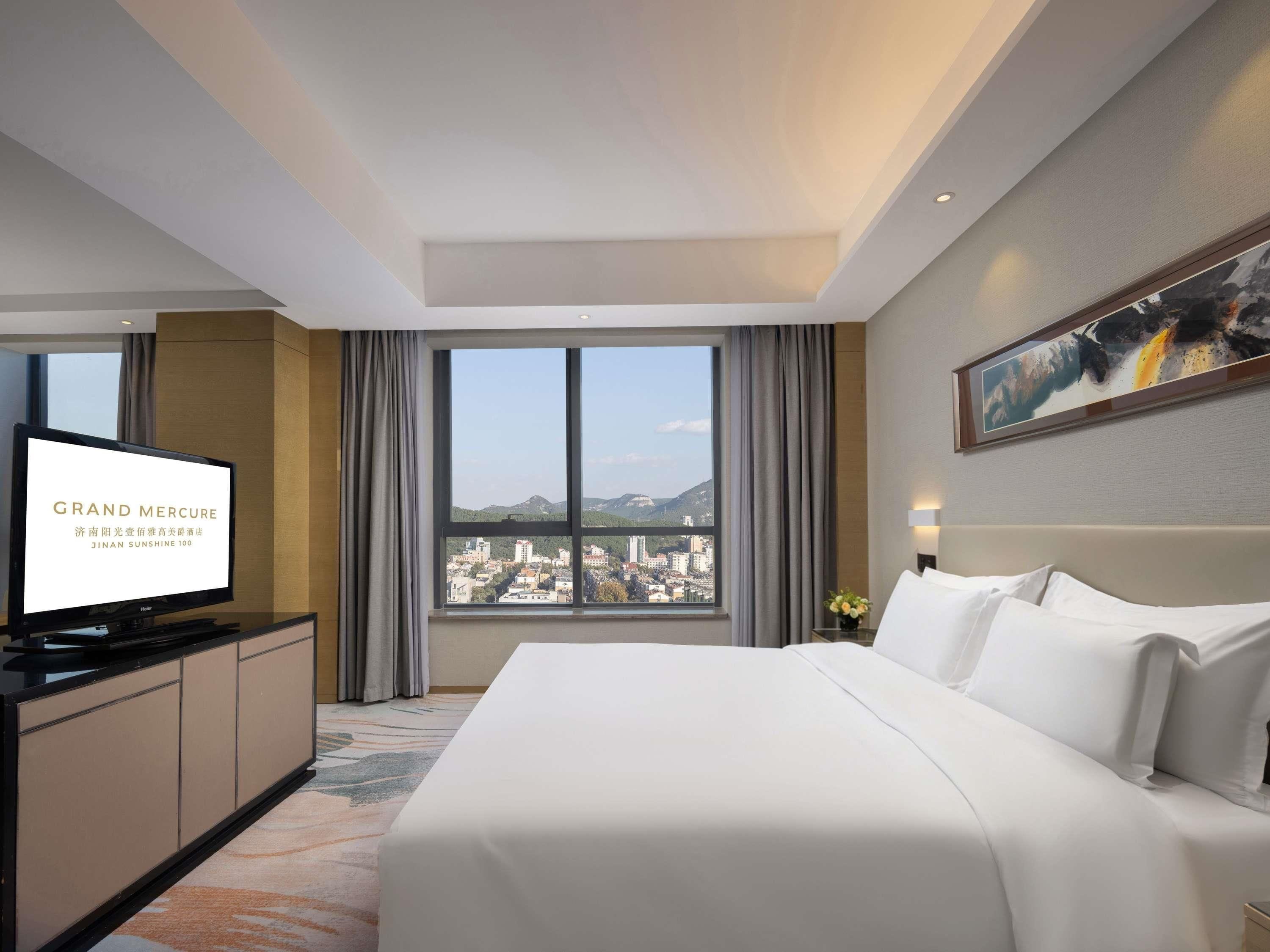 A hotel room with a bed, a TV, a desk, and a large window with a city view.