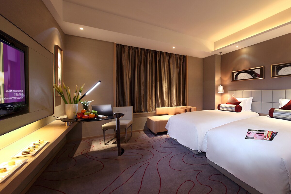 A hotel room with two beds, a desk with a laptop, a TV, and a dining area with a fruit platter.