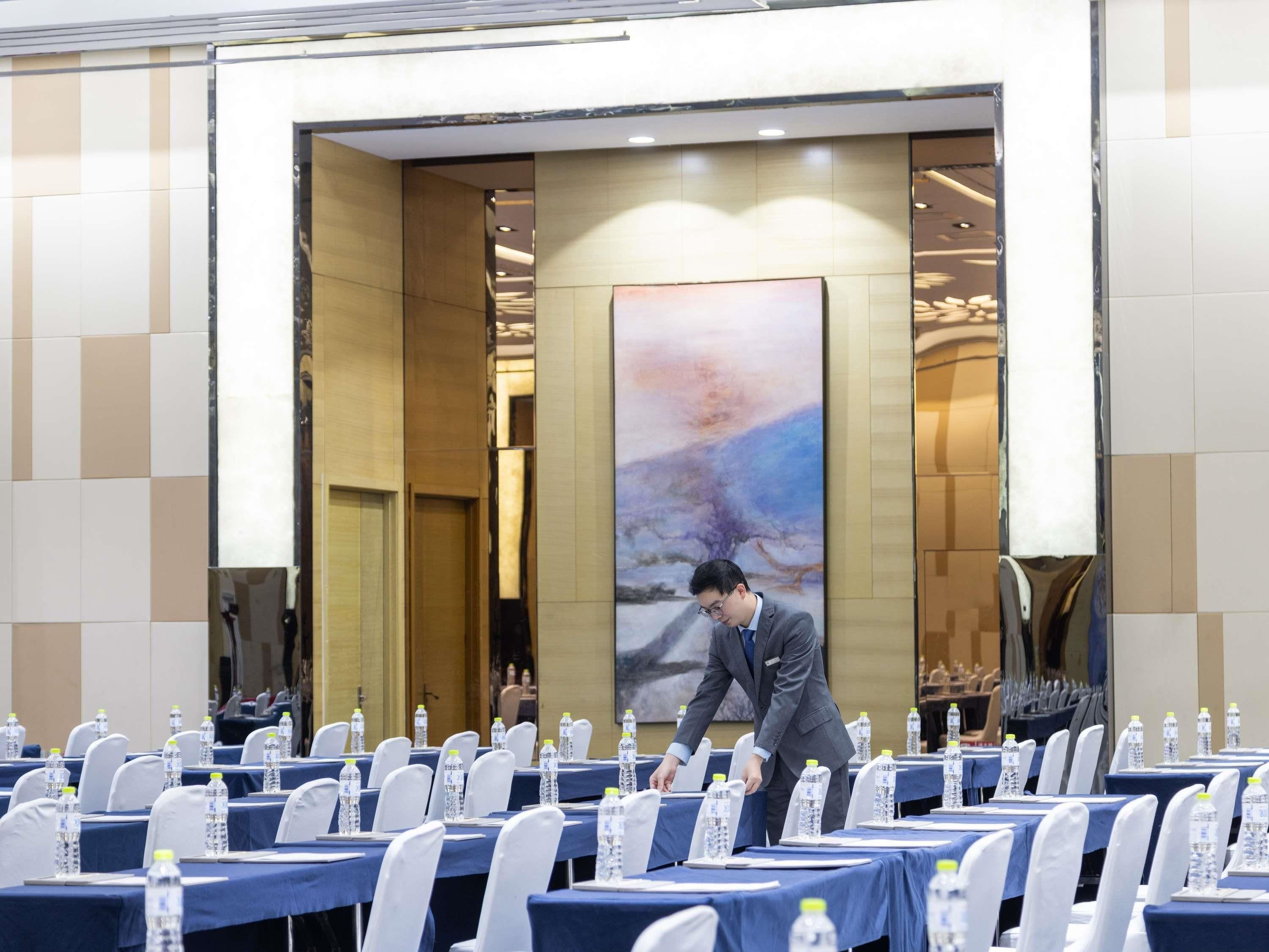 A conference room with tables set for a meeting, a large abstract painting on the wall, and a person adjusting the table settings.