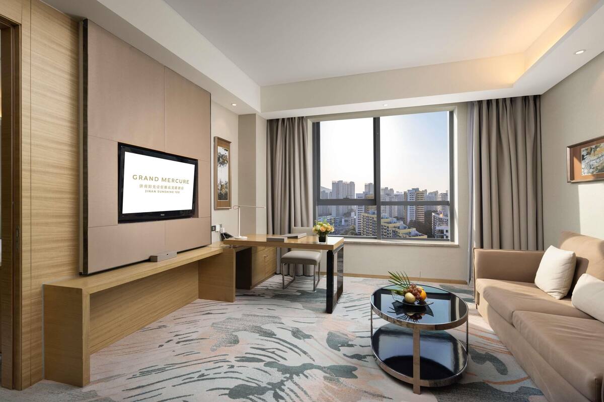 Ein modernes Hotelzimmer mit großem Fenster, einem Flachbildfernseher, einer Couch, einem Couchtisch und einem Schreibtisch mit Stuhl.