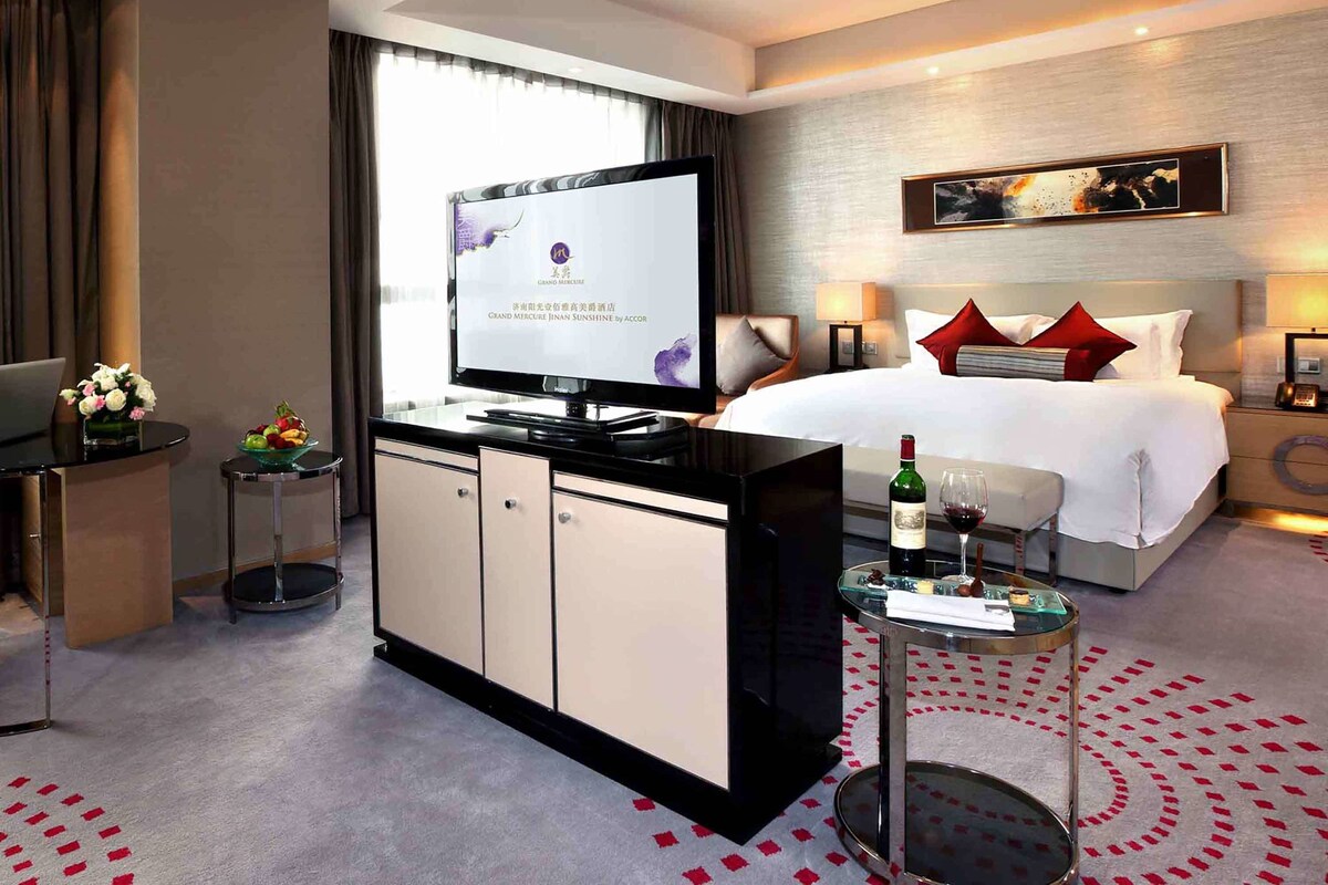 A hotel room with a bed, a TV, a desk with a computer, a small table with a bottle and glass, and a decorative wall piece.