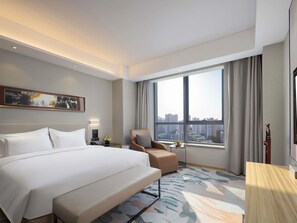 Une chambre d’hôtel avec un grand lit, une chaise, un bureau, une fenêtre donnant sur la ville et un tableau au mur.