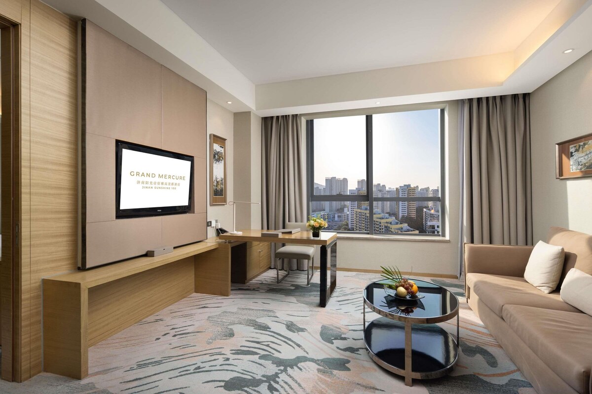 A modern hotel room with a large window, a flat-screen TV, a sofa, a coffee table, and a desk with a chair.