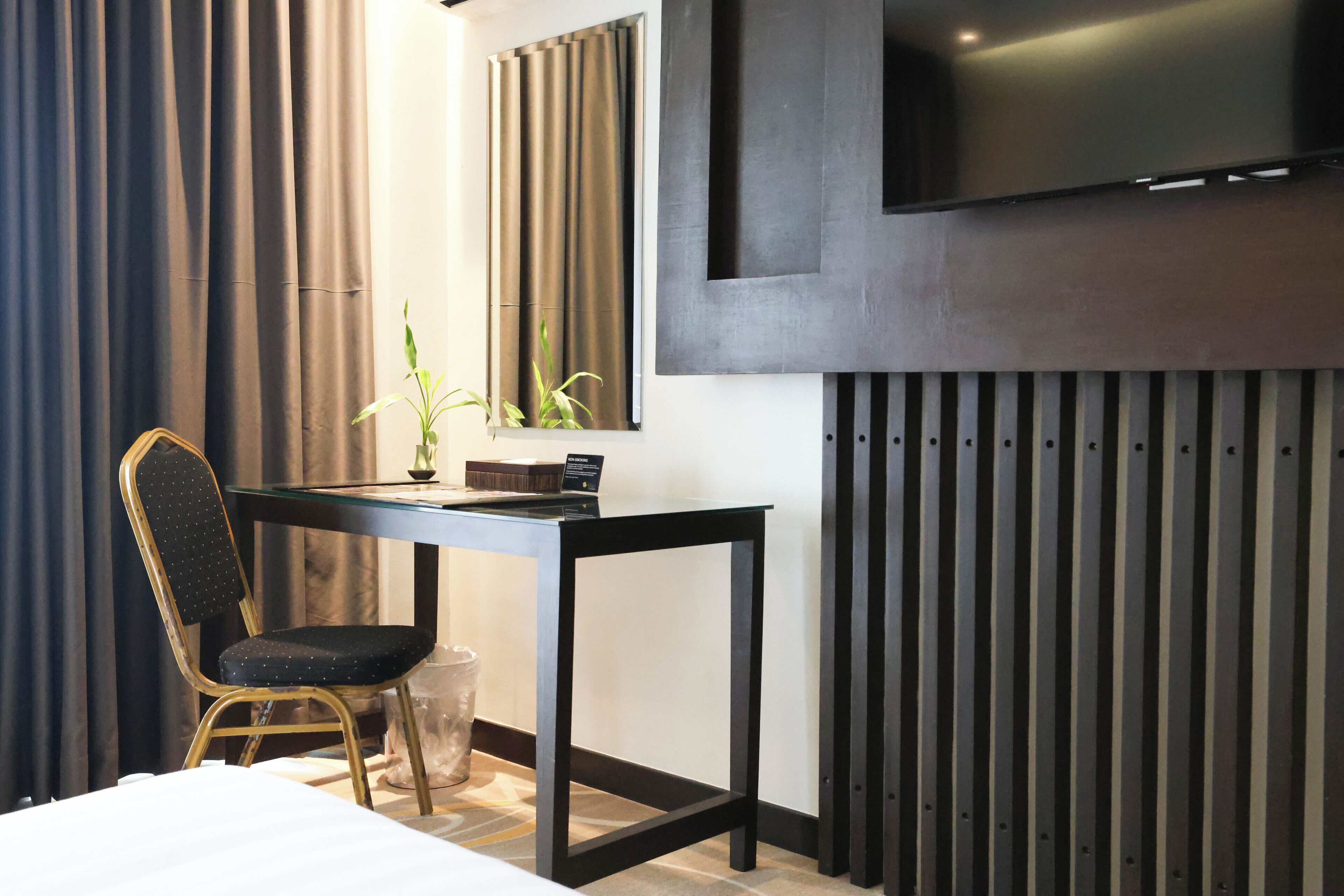A modern hotel room with a bed, a desk, a chair, a potted plant, a television, and a wall-mounted panel.