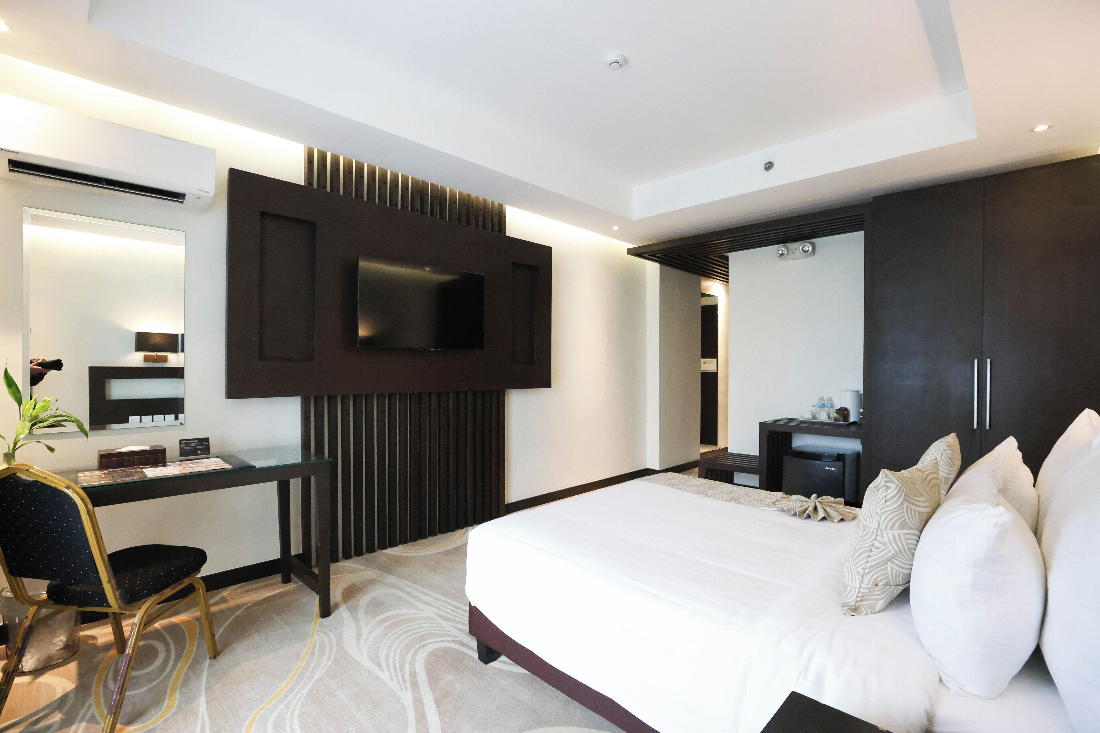 A modern hotel room with a large bed, a desk with a chair, a flat-screen TV mounted on the wall, and a dark-colored wardrobe.