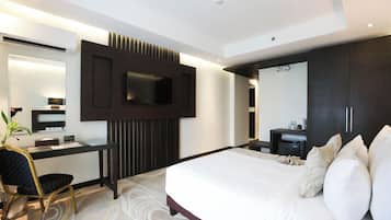 A modern hotel room with a large bed, a desk with a chair, a flat-screen TV mounted on the wall, and a dark-colored wardrobe.