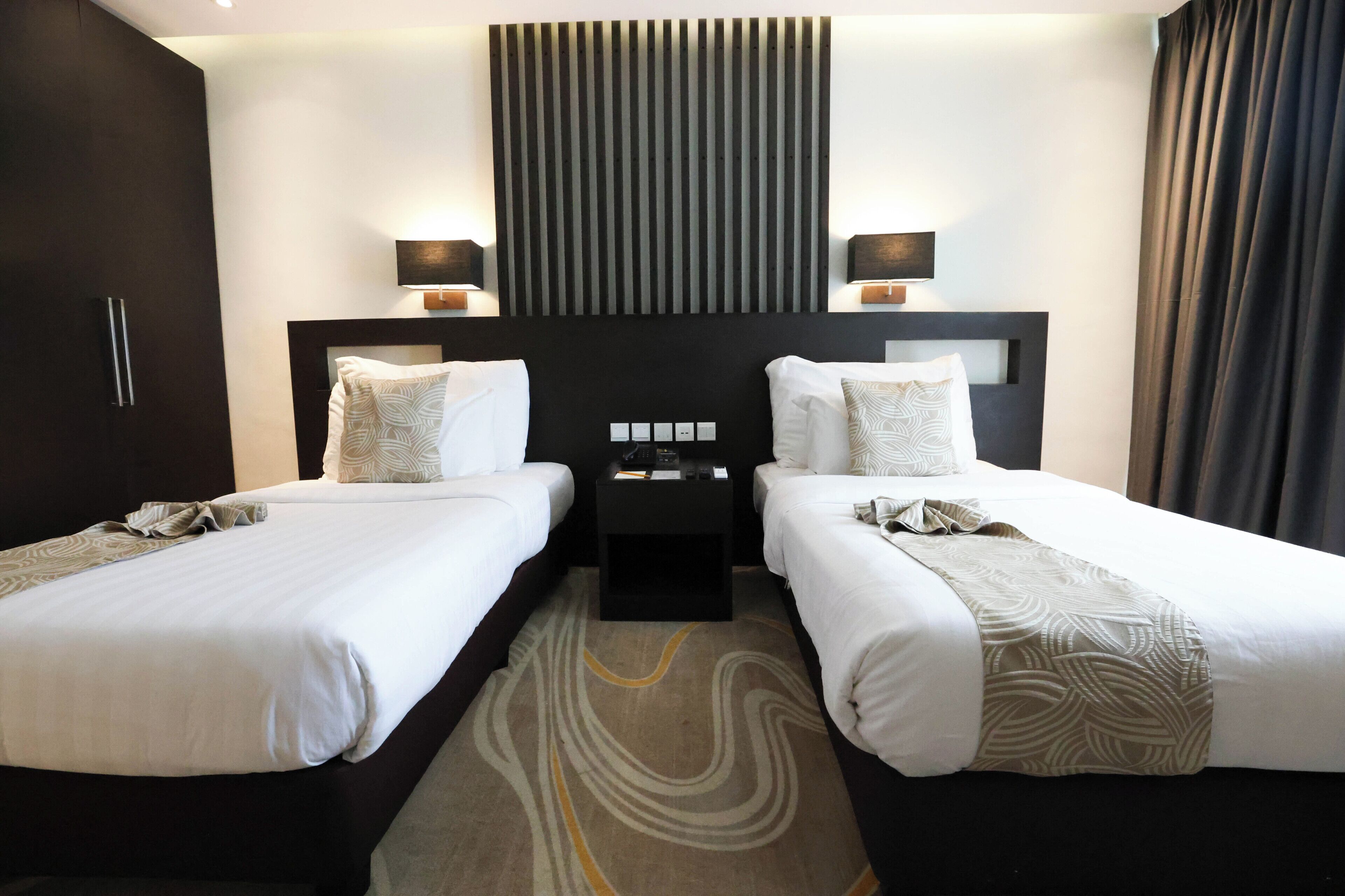 Une chambre d’hôtel avec deux lits, une tête de lit sombre, des lampes de chevet, une petite table et un tapis à motifs.