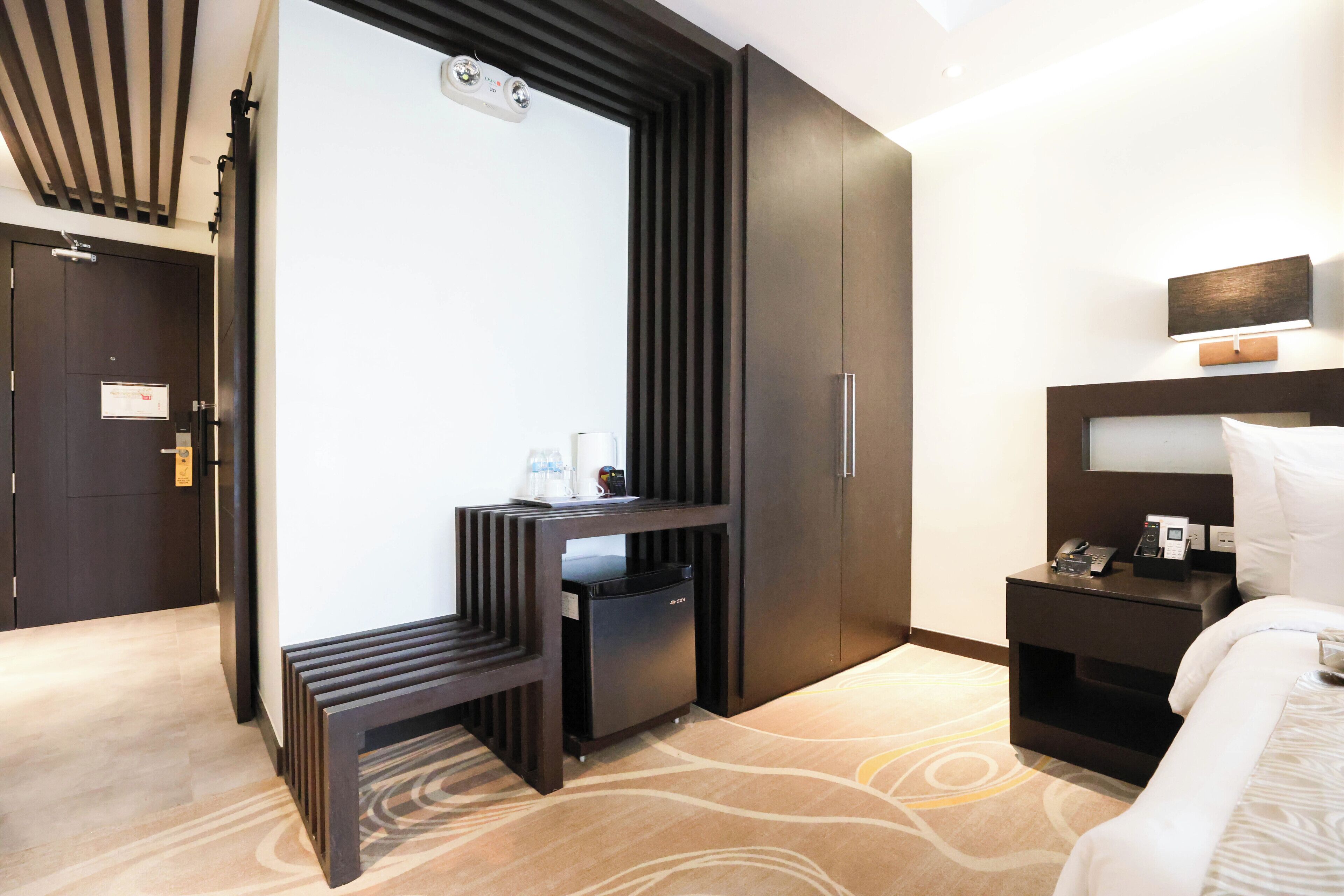A modern hotel room with a bed, bedside table, nightstand, and a built-in desk with a chair.