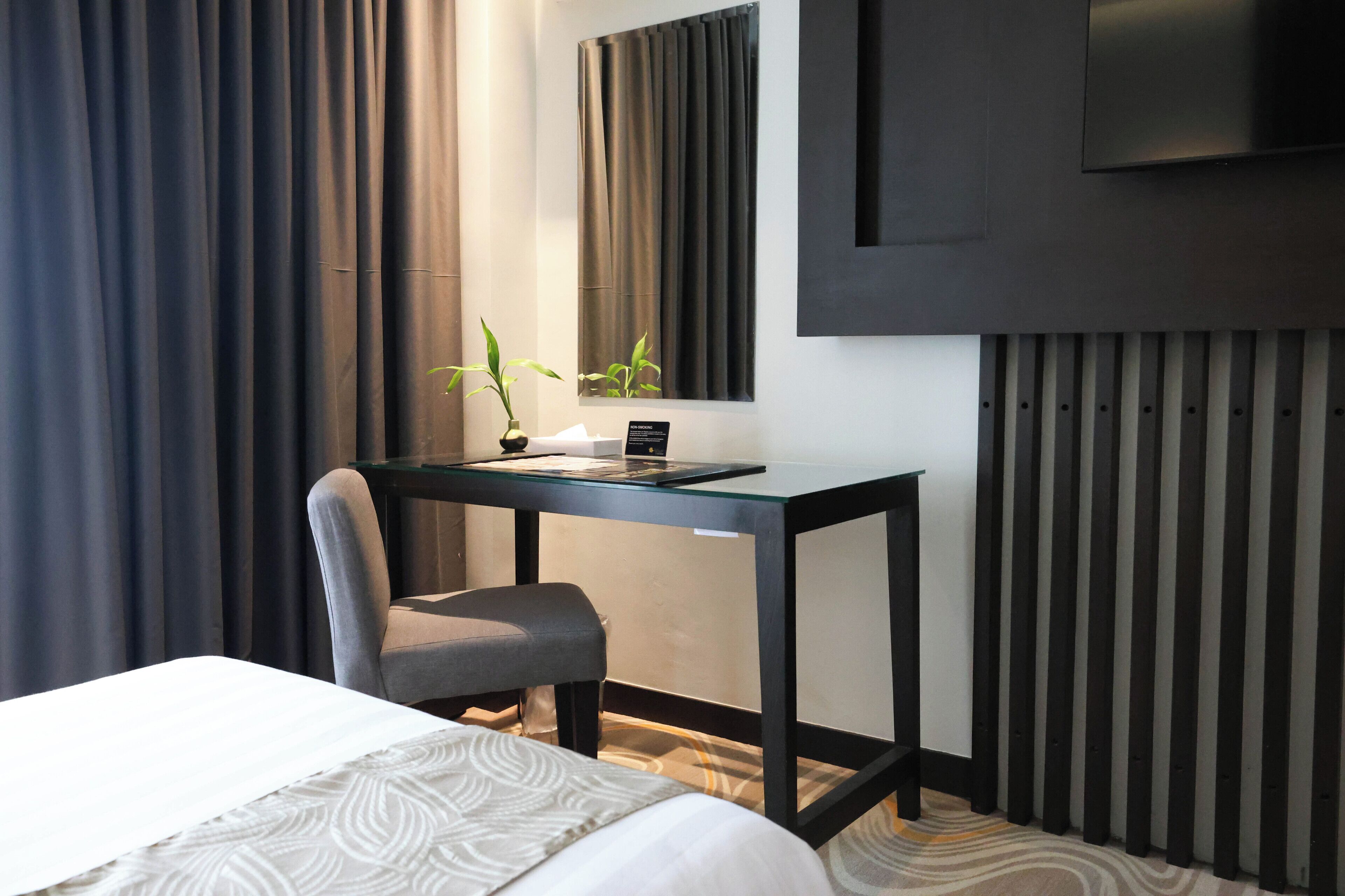 Moderni hotellihuone, jossa on sänky, työpöytä tietokoneella, tuoli ja ruukkukasvi.