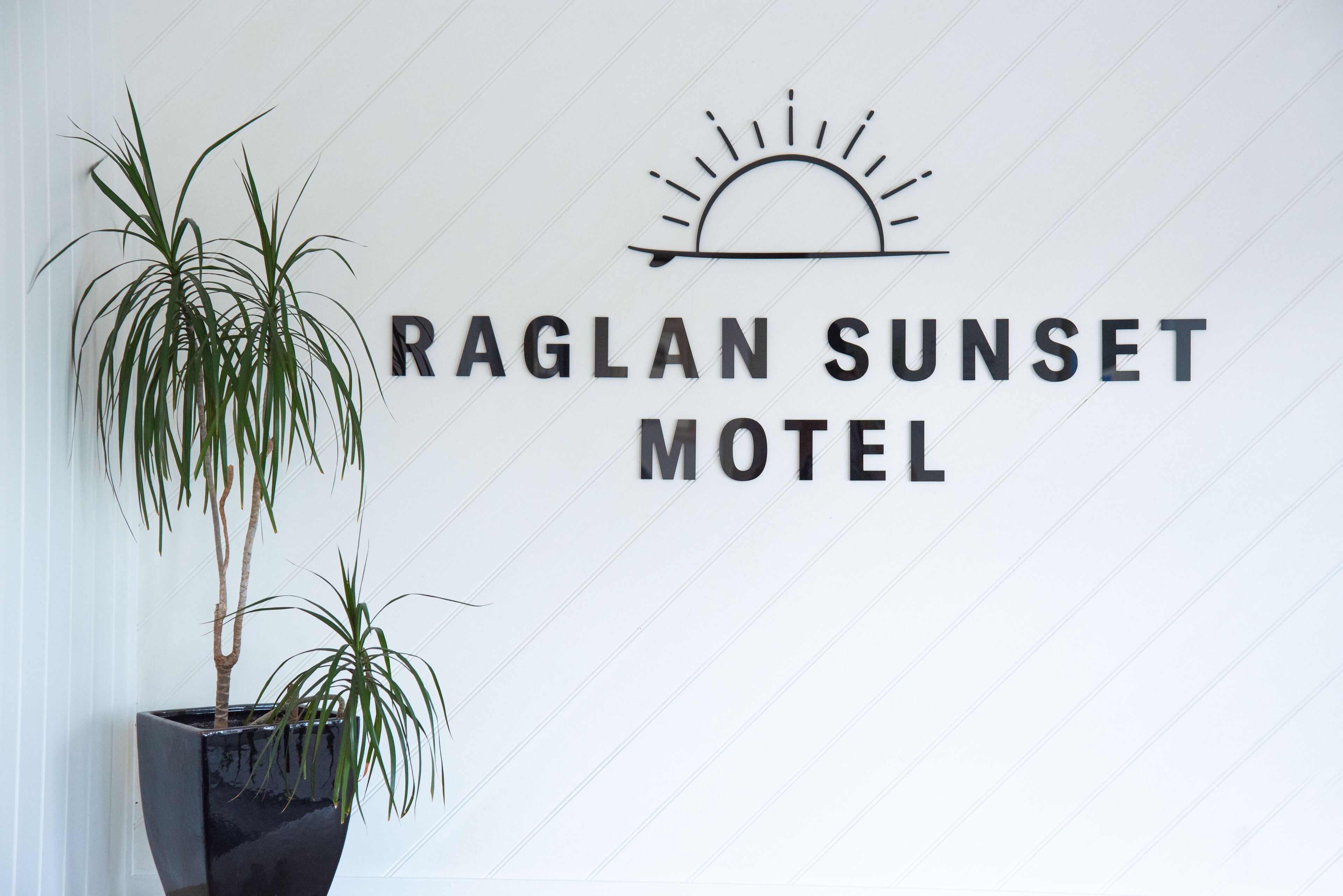 Una pared con el texto "RAGLAN SUNSET MOTEL" y un gráfico de sol. A la izquierda, hay dos plantas en macetas.