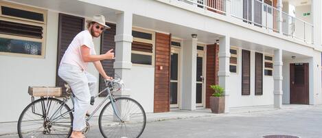 A person on a bicycle outside a building with a balcony, a wooden door, and a potted plant.