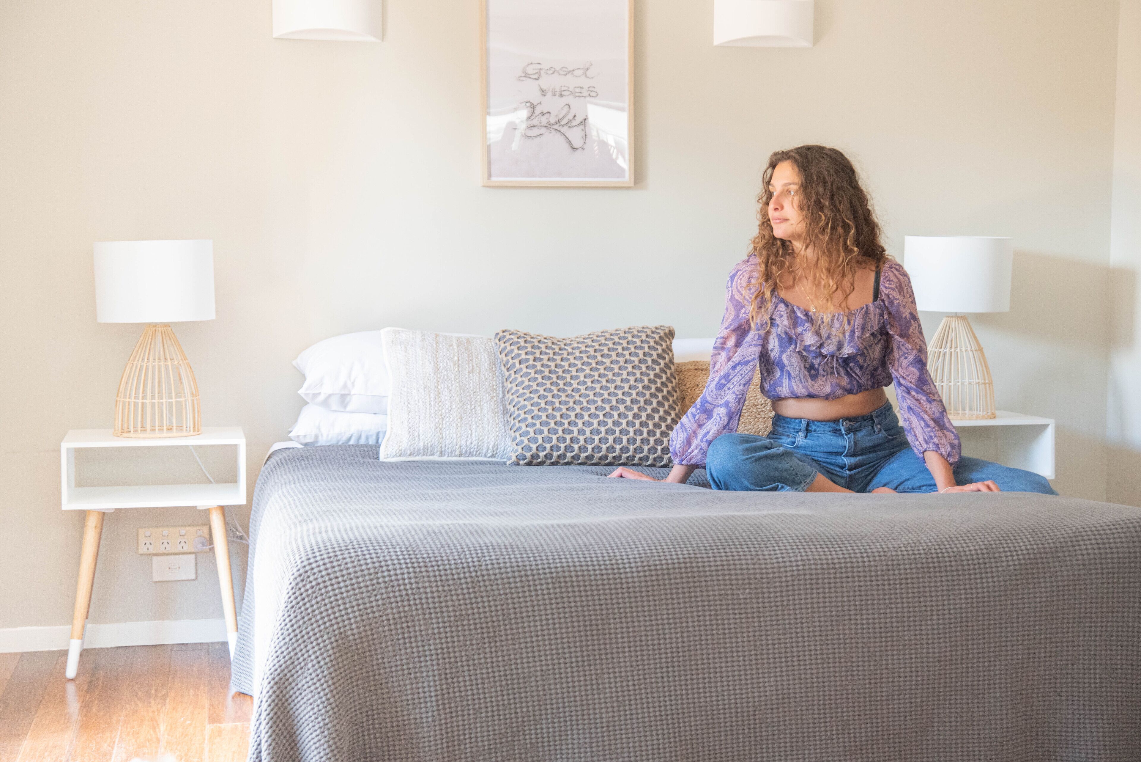 Une personne est assise sur un lit dans une pièce dont le mur est blanc. On y voit un cadre avec une citation et deux tables de chevet avec des lampes.