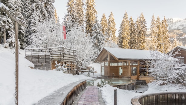 A wooden cabin with a glass enclosure, surrounded by snow-covered trees and a wooden deck leading to a hot tub. - Hotel Bad Schoergau (Sarentino)