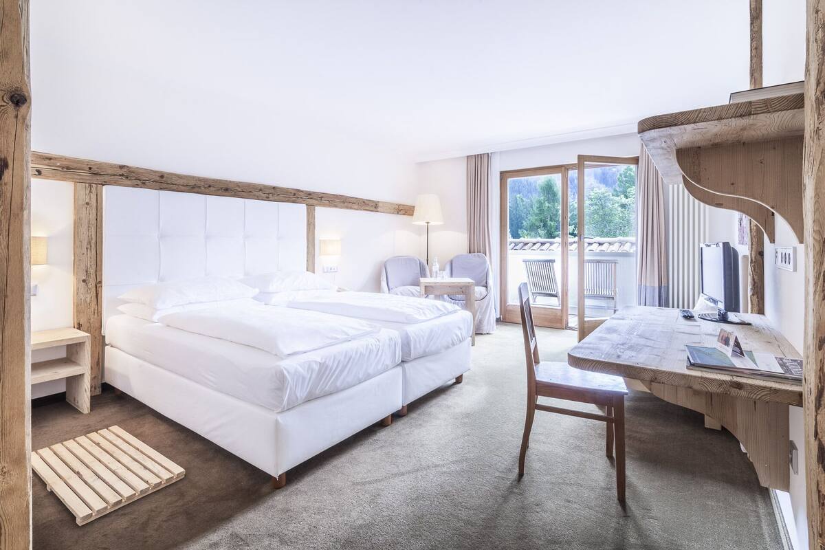 A hotel room with a large bed, a desk, a chair, and a balcony with a view of greenery.