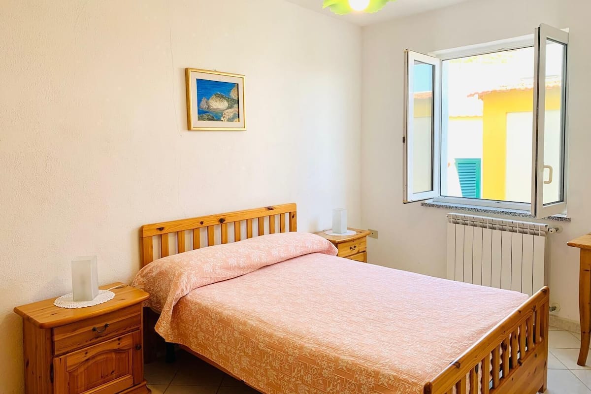 A bedroom with a wooden bed, a nightstand, a bedside table, a window with an open shutter, a radiator, and a framed picture on the wall.
