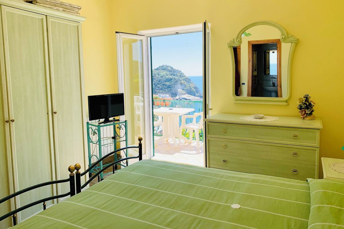 A bedroom with a bed, a dresser, a television, and a balcony with a view of the sea.