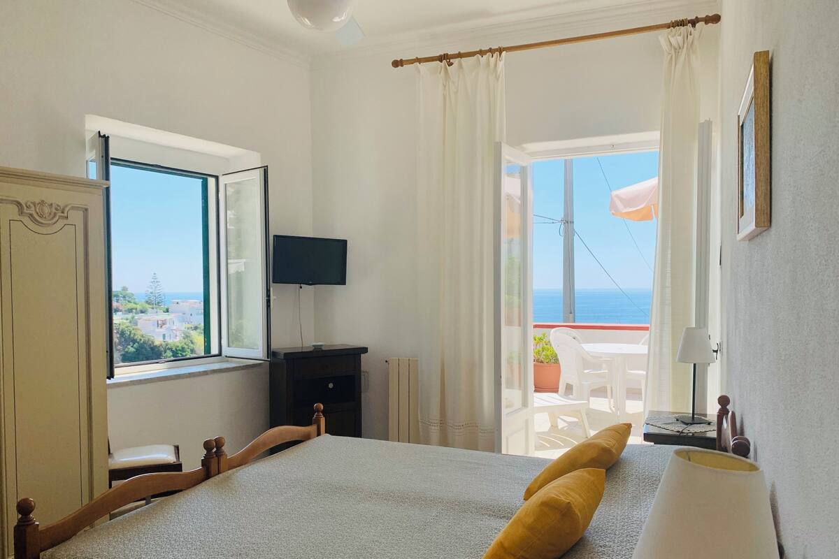 A bedroom with a bed, a dresser, a TV, and a balcony with a view of the sea.