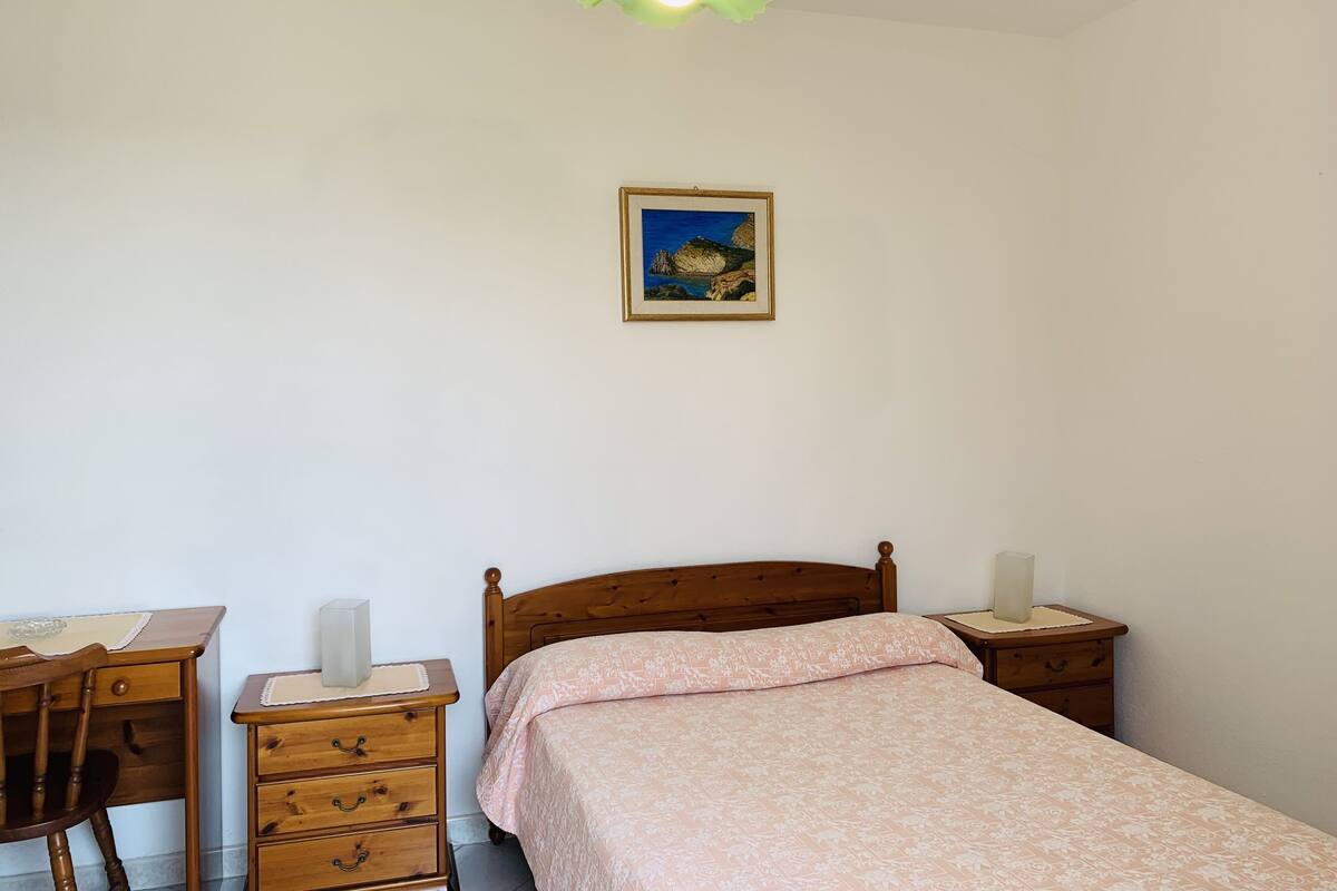 A bedroom with a wooden bed, a desk, a chair, a dresser, a bedside table, and a framed picture on the wall.