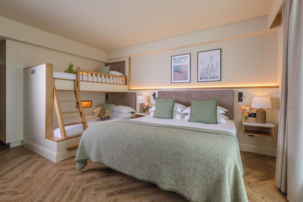 A hotel room with a bunk bed, a desk, two bedside lamps, and a bed with a green blanket.