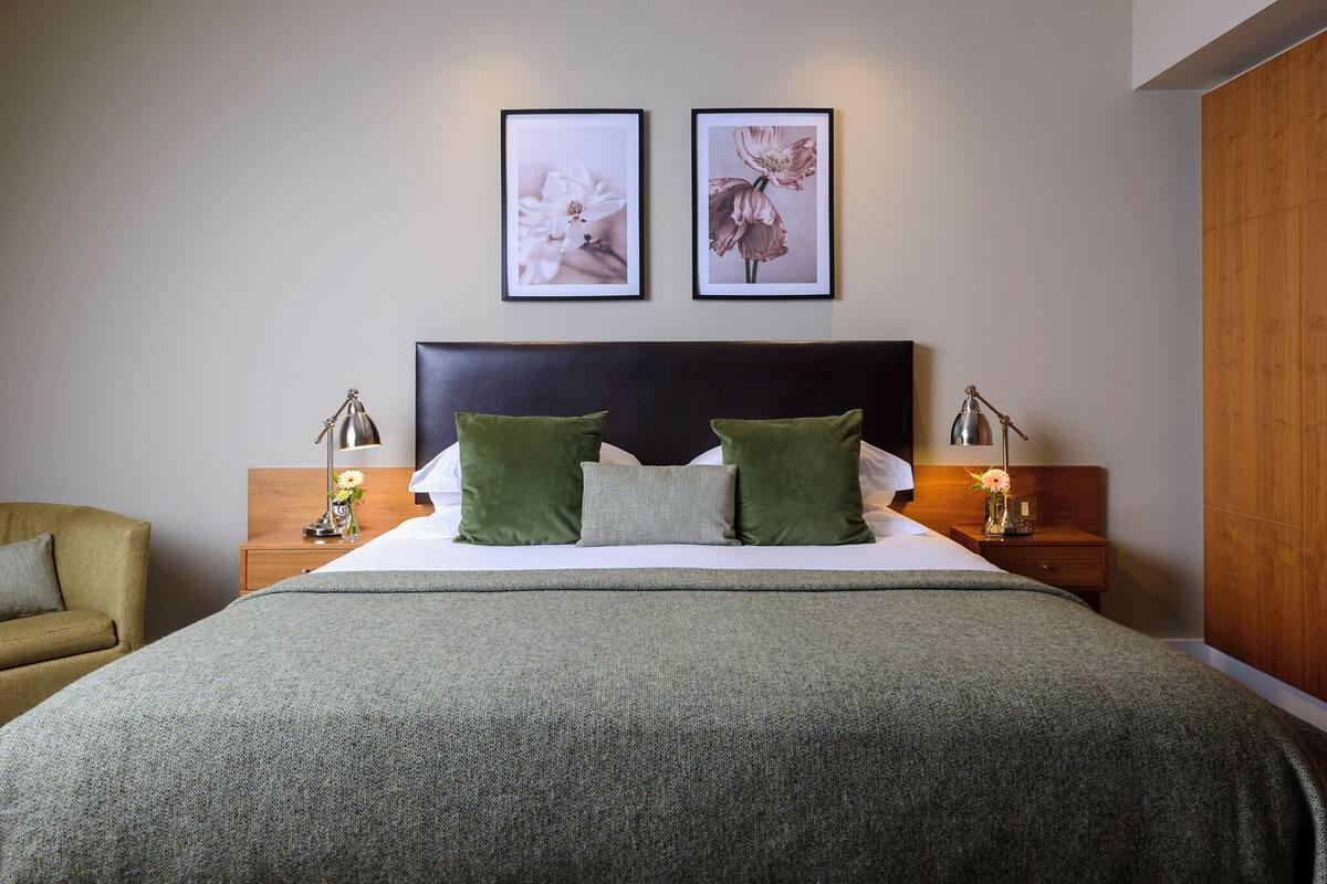 Ein ordentlich eingerichtetes Schlafzimmer mit einem großen Bett, grünen Kissen, zwei gerahmten floralen Bildern an der Wand und Nachttischlampen.