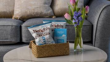 A wicker basket with popcorn bags and a milk chocolate bar on a white table, with a vase of flowers and a grey sofa in the background.