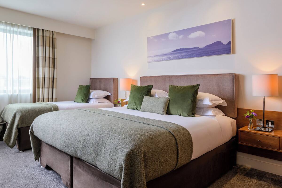 Ein Hotelzimmer mit zwei Betten, jeweils mit grüner Bettwäsche, einem Nachttisch mit Lampe und einem Landschaftsbild an der Wand.