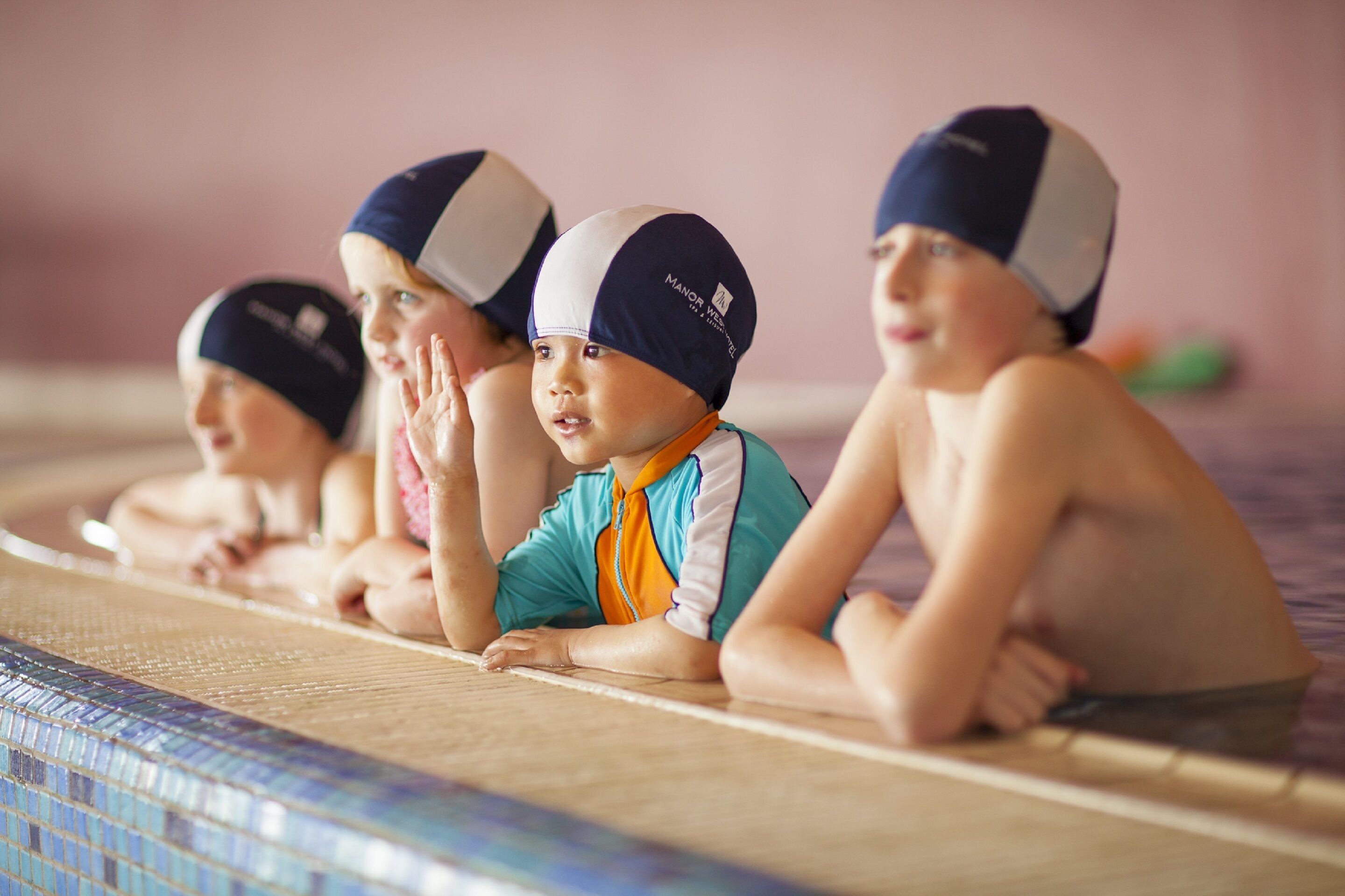 A group of children sitting by a poolside, wearing swim caps and goggles.