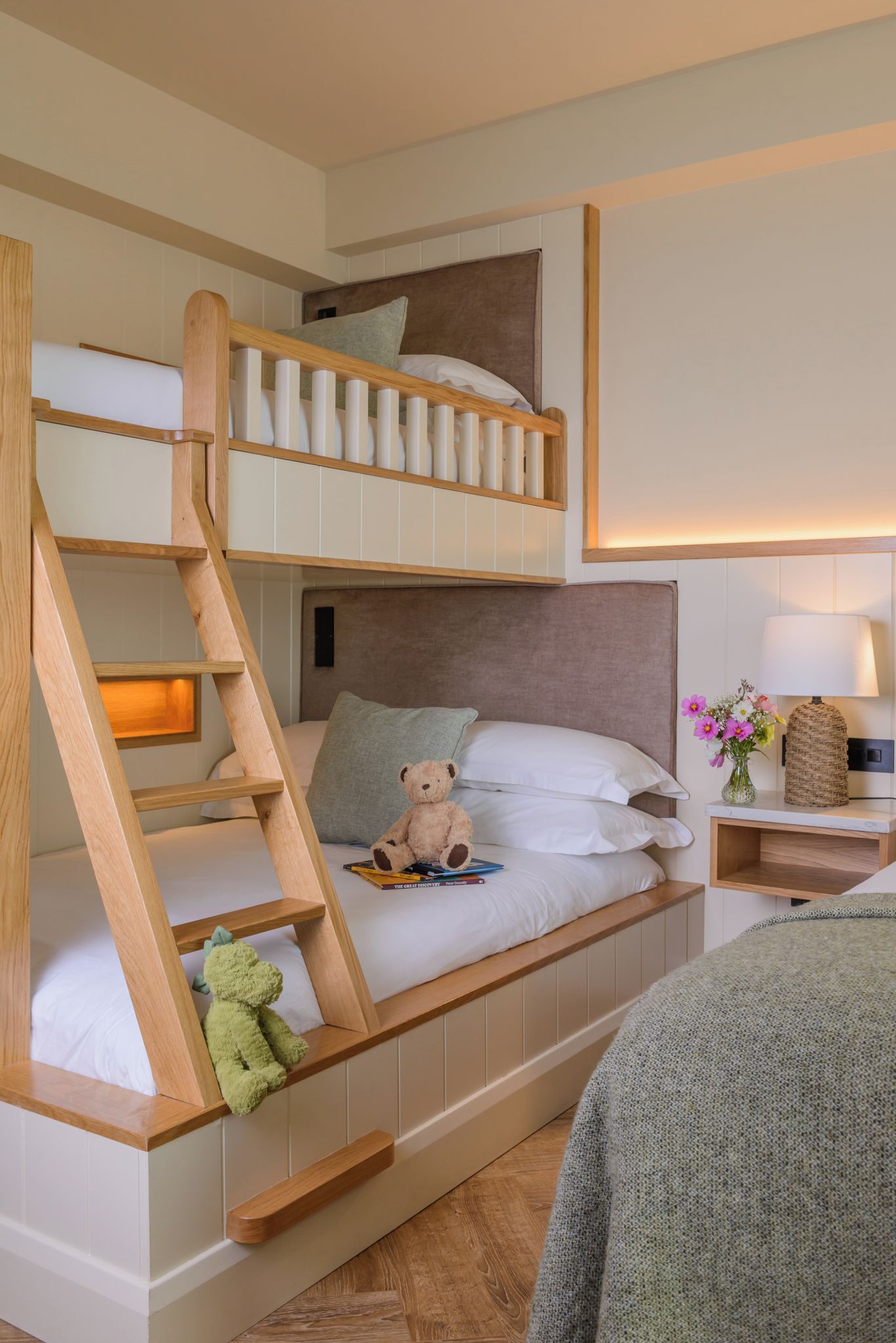 Een stapelbed met een houten trapje, een nachtkastje met een lamp, en een teddybeer op het onderste bed.