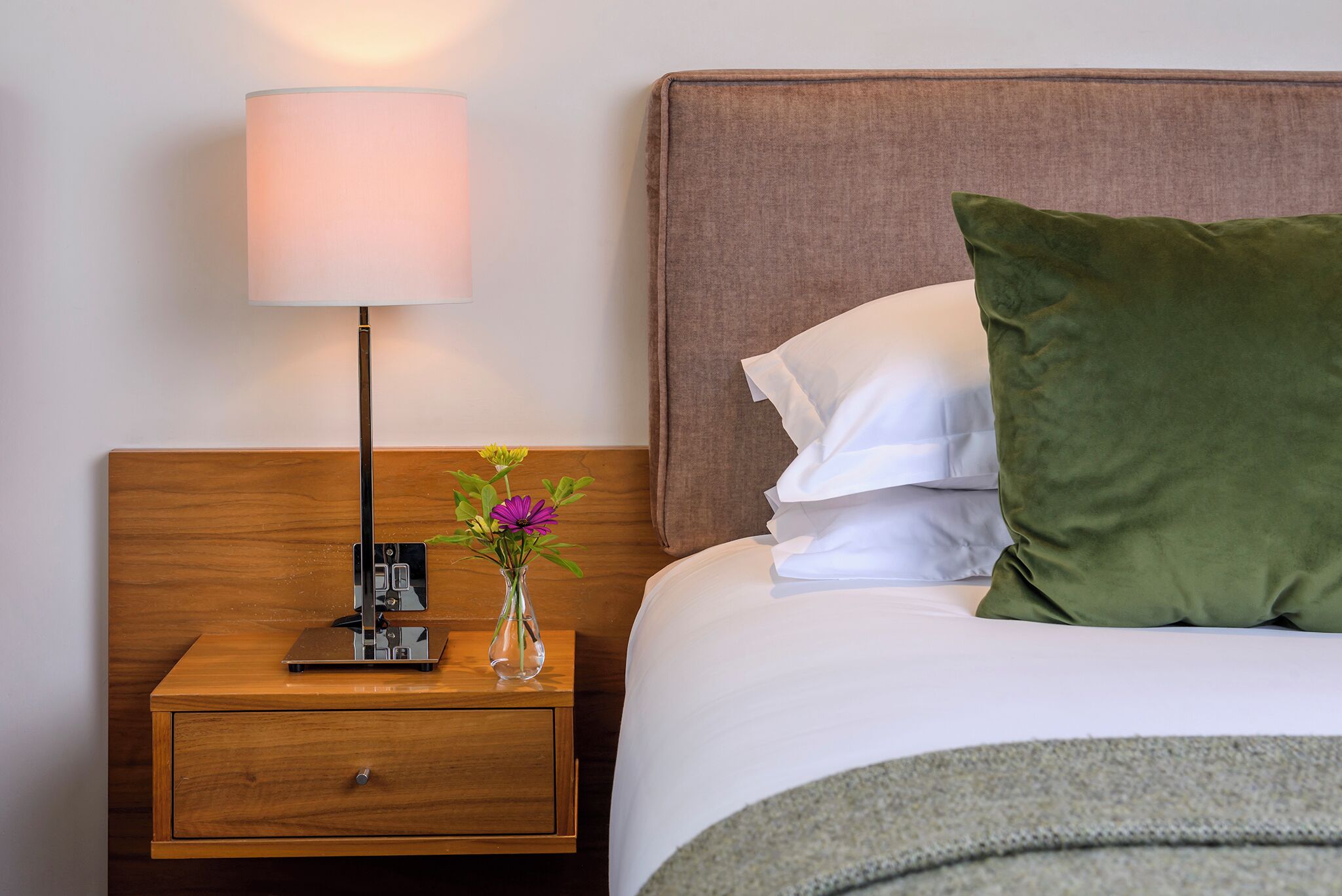 Uma cama bem arrumada com lençóis brancos, um travesseiro verde e uma mesa de cabeceira com abajur e um vaso de flores.