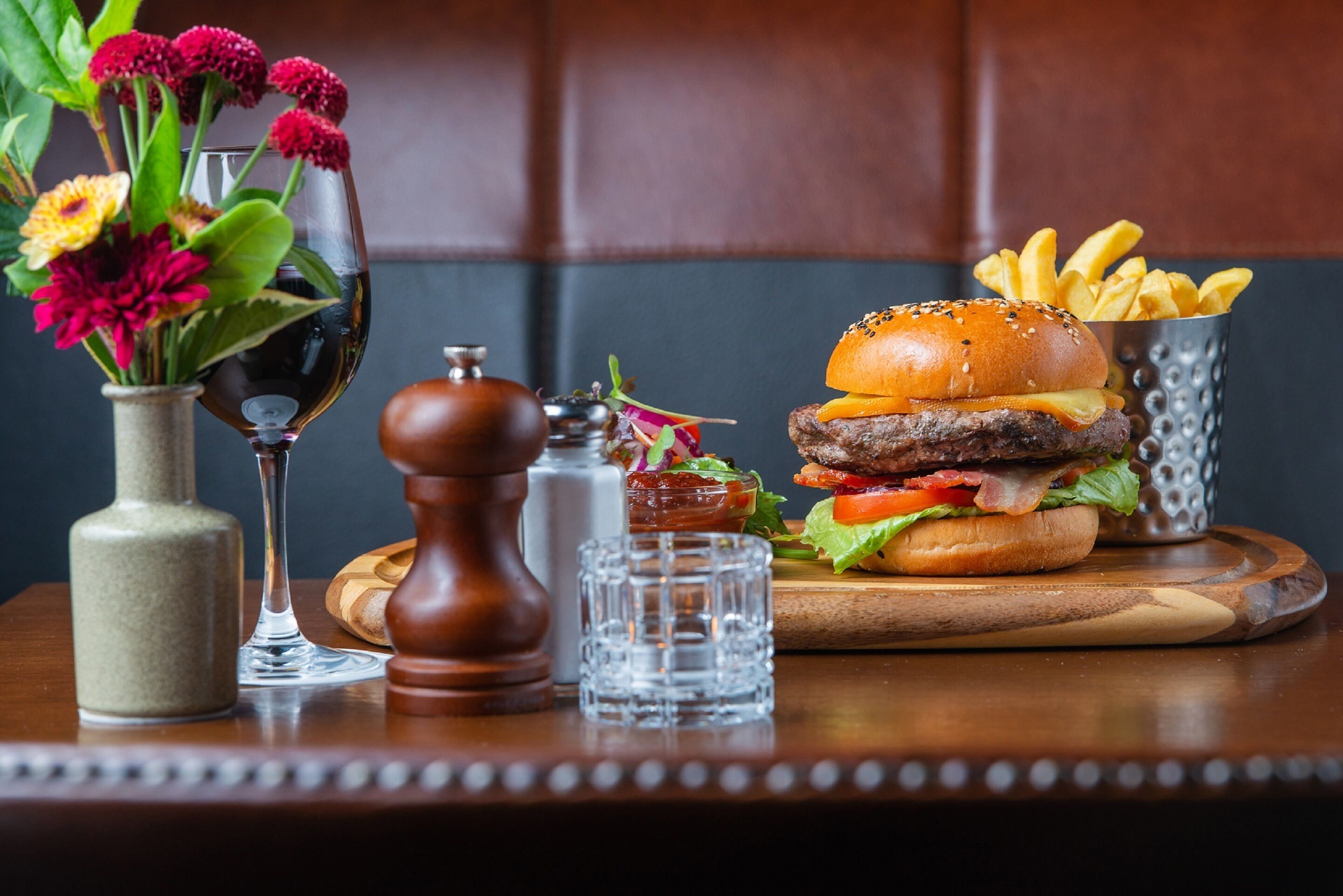 A wooden table with a burger, fries, a glass of wine, and a vase with flowers.