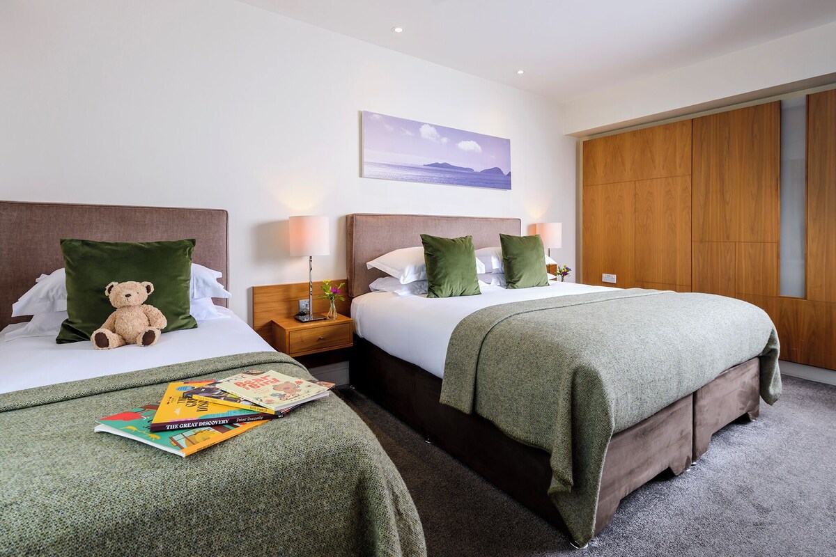 A hotel room with two beds, green bedding, a teddy bear on one bed, a nightstand with a lamp, a painting on the wall, and a wooden wardrobe.