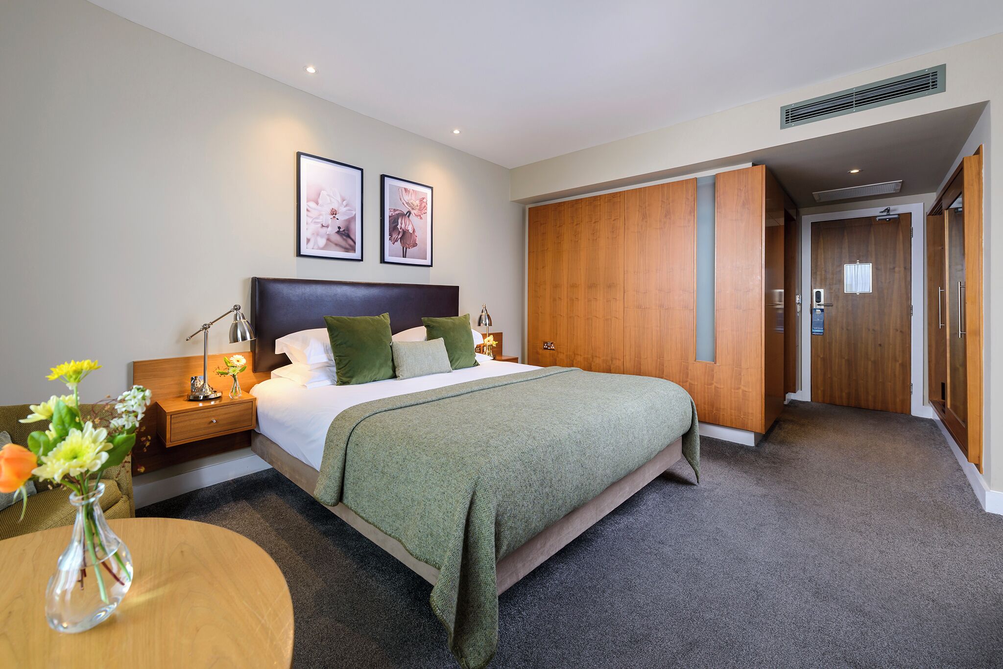 Habitación de hotel con cama, mesitas de noche, un armario de madera y una pequeña mesa con un florero.