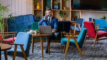 A man in a blue suit sitting on a blue sofa with a laptop, surrounded by colorful chairs and a TV in the background.
