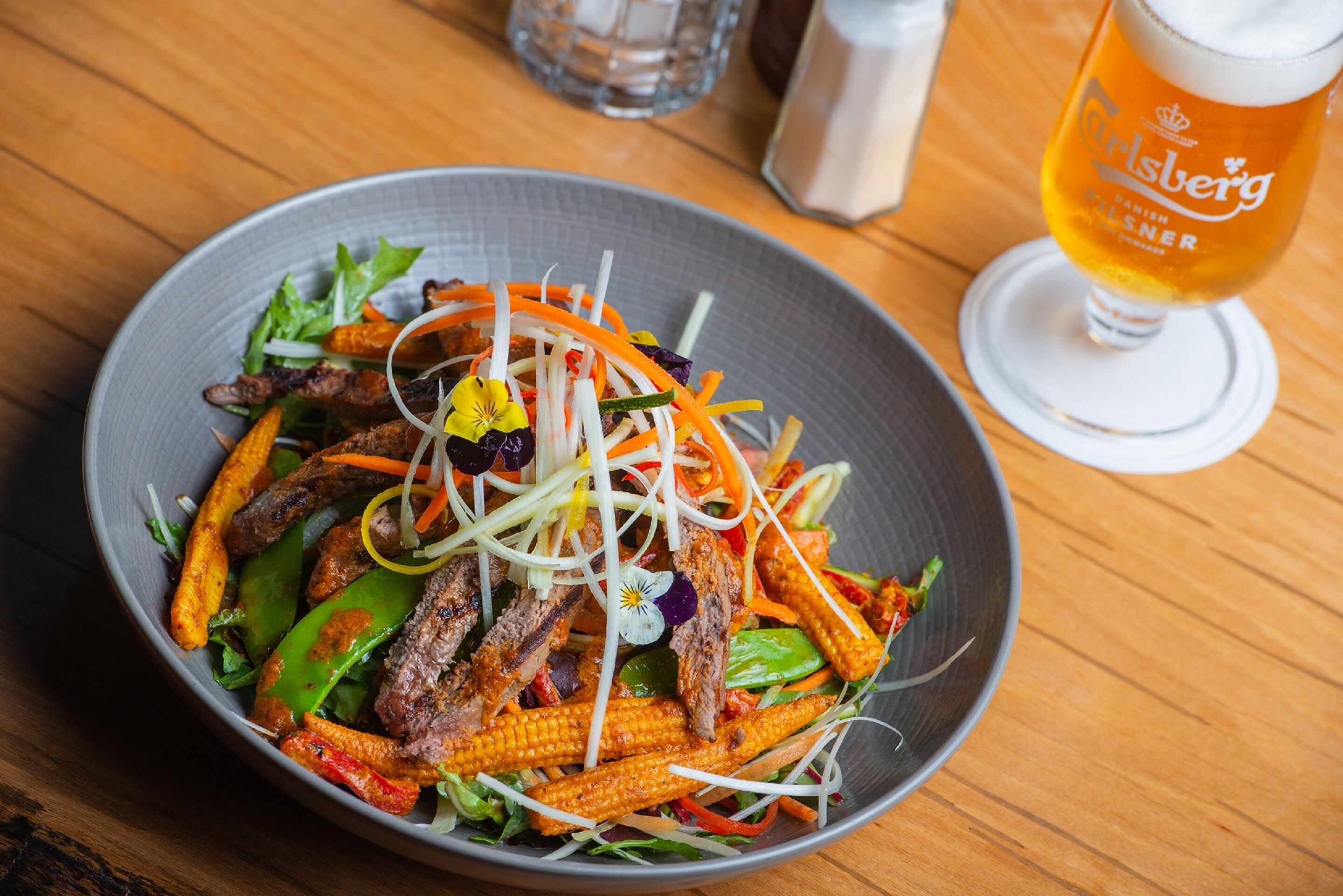Un bol de ensalada con carne y verduras, una jarra de cerveza y saleros y pimenteros sobre una mesa de madera.