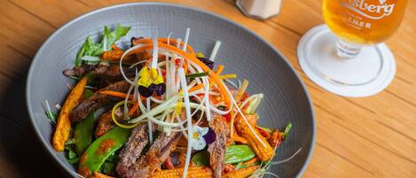 A bowl of salad with meat and vegetables, a glass of beer, and salt and pepper shakers on a wooden table.