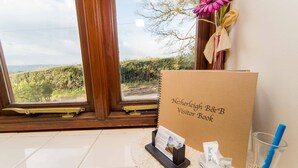 Un atril para libro de visitas con un folleto, un sujetapapeles y un vaso con pajita sobre un suelo de baldosas cerca de una ventana que ofrece vistas a la vegetación.