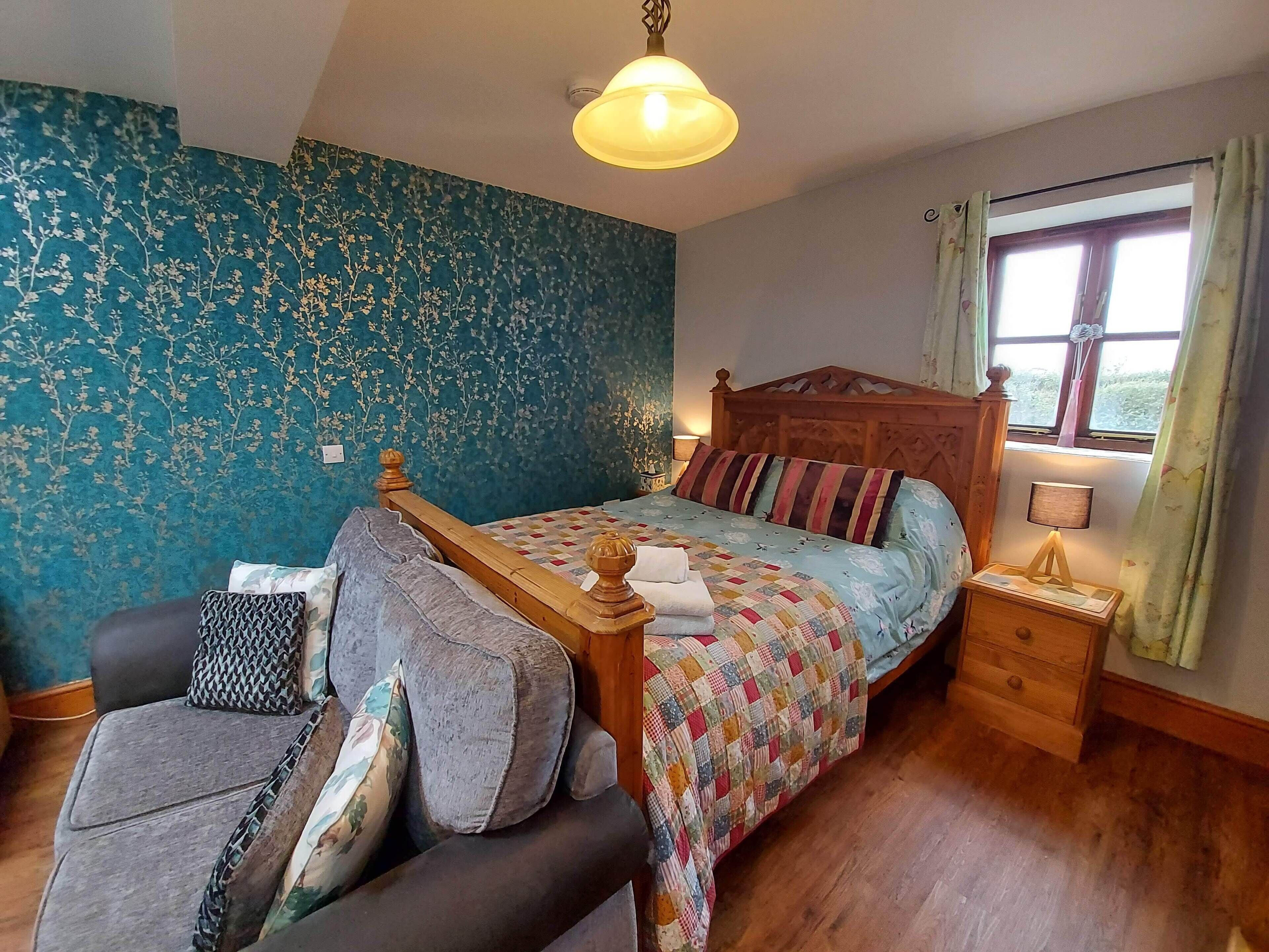 Un dormitorio con una cama de madera, una pared con estampado floral azul, una ventana con cortinas, una mesita de noche y un sofá con cojines.