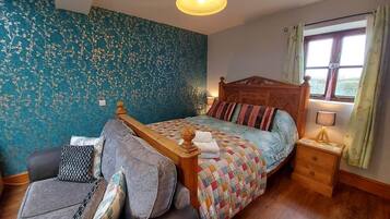 Un dormitorio con una cama de madera, una pared con estampado floral azul, una ventana con cortinas, una mesita de noche y un sofá con cojines.