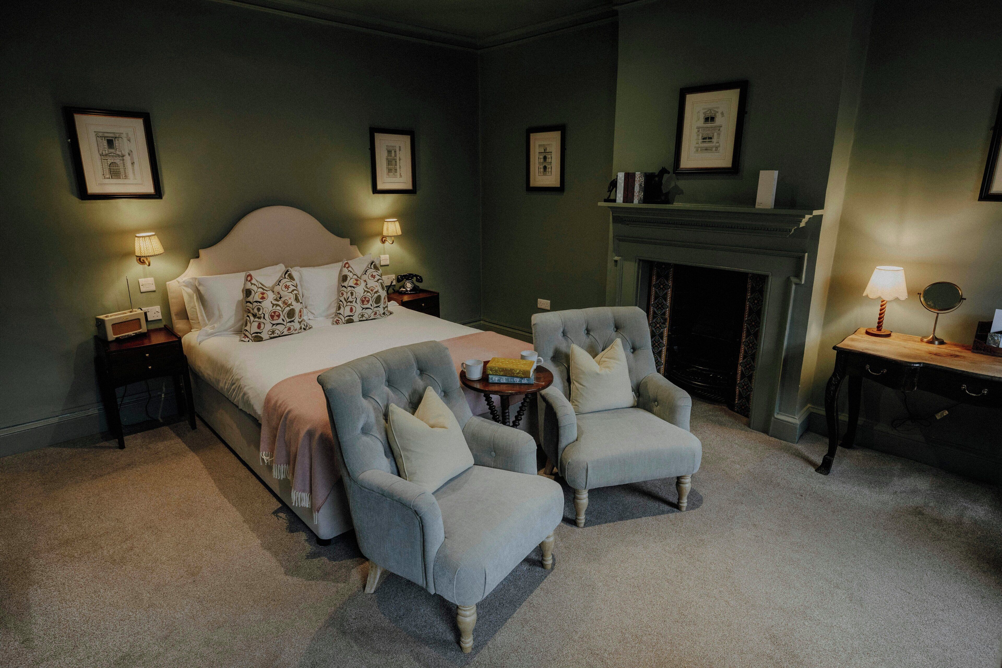 A bedroom with a large bed, two armchairs, a small table, a fireplace, and framed pictures on the walls.