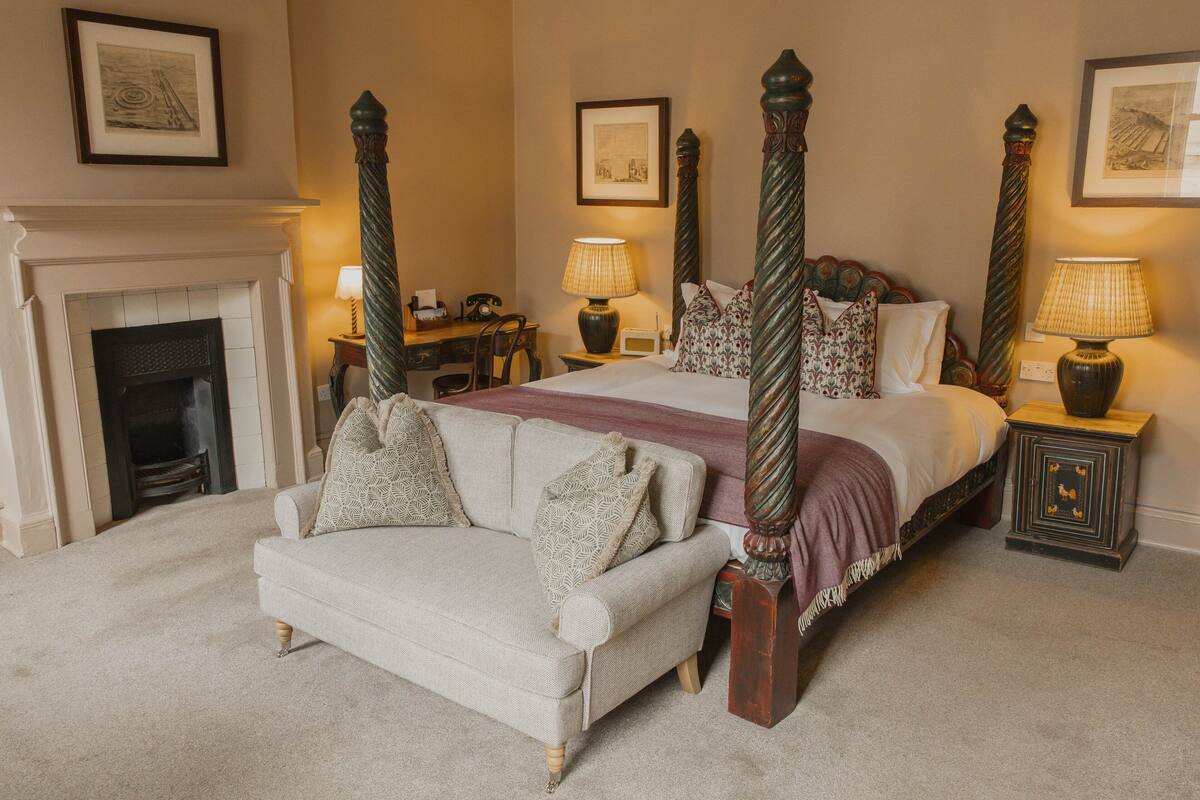 A four-poster bed with a headboard, a fireplace, a sofa, and two nightstands with lamps.
