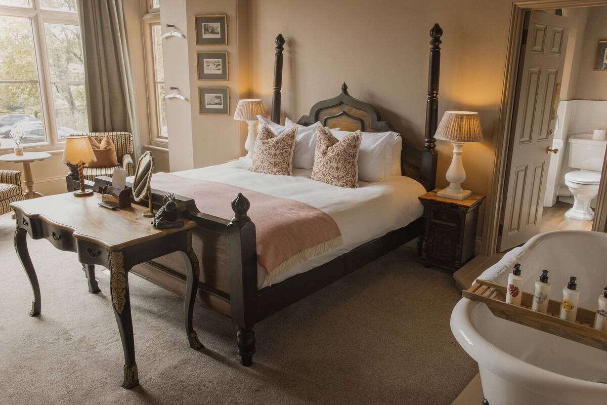 A bedroom with a four-poster bed, bedside tables, a vanity with a mirror, and a bathtub.