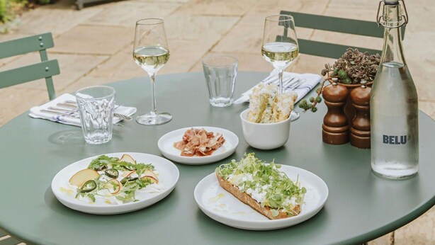 Un área de comedor al aire libre con una mesa preparada para dos personas, con platos de comida, copas de vino y una botella de agua Belu.