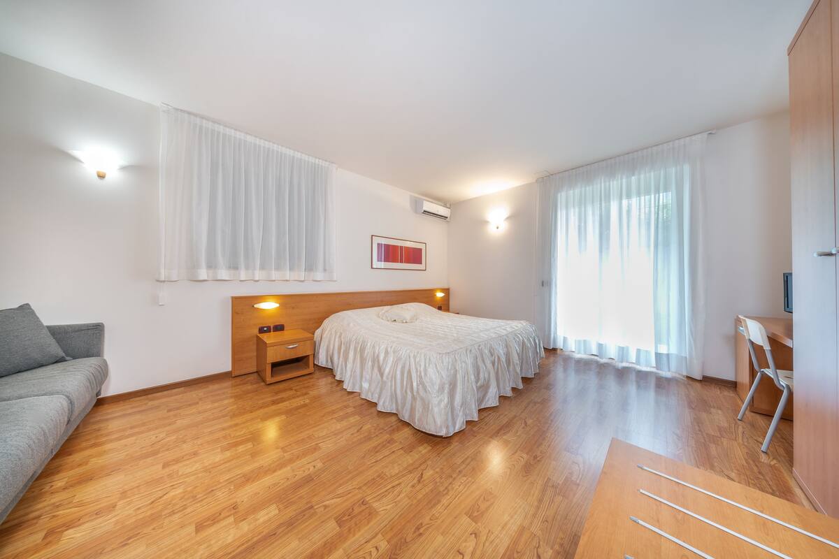 A modern bedroom with a wooden floor, a bed with a white bedspread, a grey sofa, a wooden nightstand, and a desk with a chair.