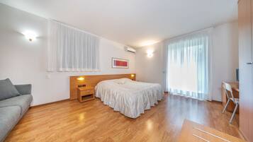 Un dormitorio moderno con piso de madera, una cama con cubrecama blanco, un sofá gris, una mesita de noche de madera y un escritorio con una silla.