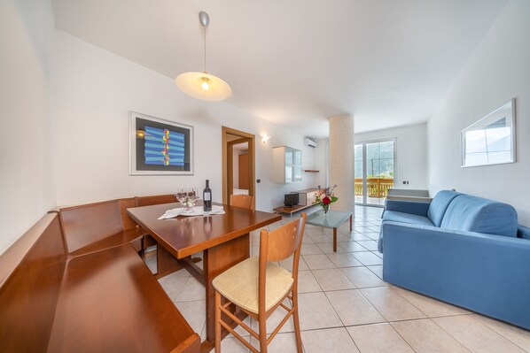 A modern living room with a wooden dining table, a blue sofa, and a large window offering a view of the outdoors.
