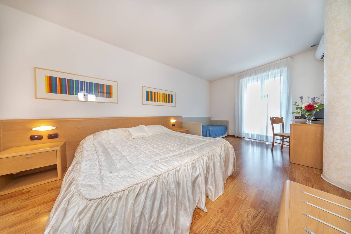 A hotel room with a large bed, wooden headboard, bedside tables, a desk, and a chair. There are two framed artworks on the wall, a window with curtains, and a small table with a vase of flowers.